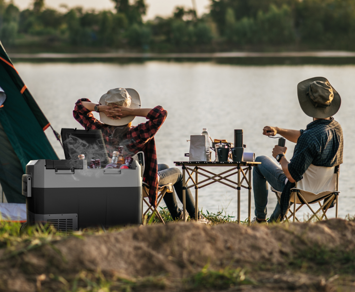 A couple relaxing in camping chairs by a lake next to an open car refrigerator filled with chilled drinks.