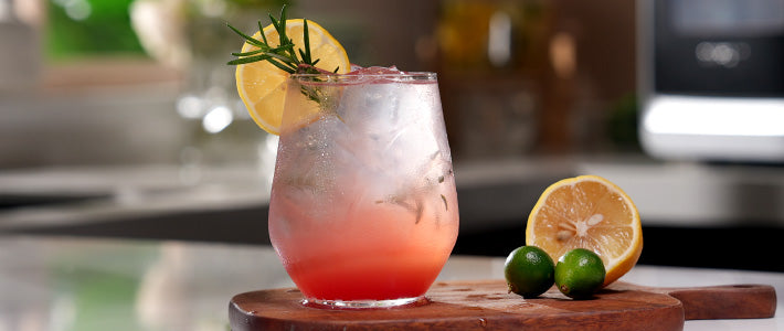 Watermelon Spark drink with LunaArc™ crescent ice, garnished with lemon and rosemary. Served on a wooden board with extra lemon and limes.