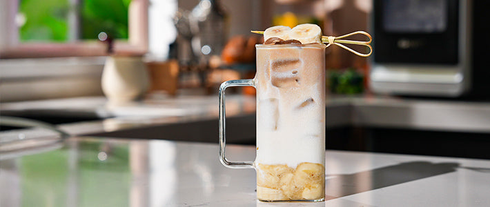 Banana Iced Latte with visible ice cubes, layered with banana, milk, and coffee, garnished with skewered banana slices.