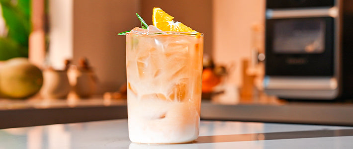 Finished orange latte in a tall glass with ice, garnished with an orange slice and rosemary sprig.