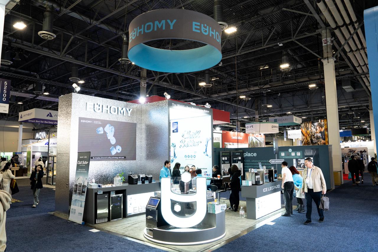 A modern indoor CES booth for Euhomy featuring a prominent blue hanging circular sign, silver textured walls, and attendees browsing the latest smart ice maker models.